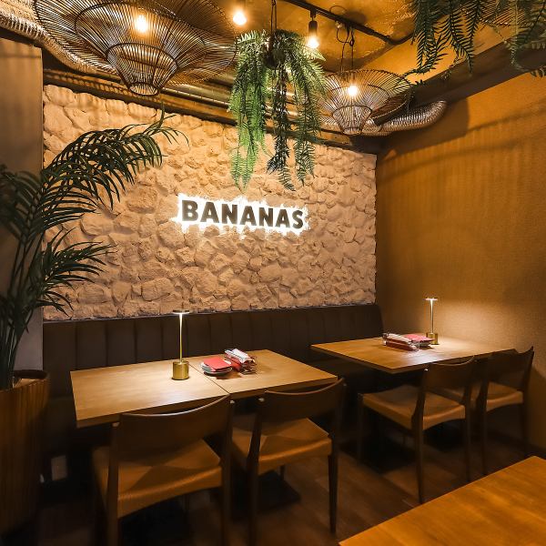 【内観】天井から吊るされたグリーンや、編み込みデザインのペンダントライトが、まさに南国リゾートのムードを演出。石垣調の壁を照らすネオンサイン「BANANAS」も、お洒落な写真映えスポットです。深みのあるモスグリーンソファと木目調のインテリアが、都会の喧騒を忘れさせる特別な空間を作り出しています。