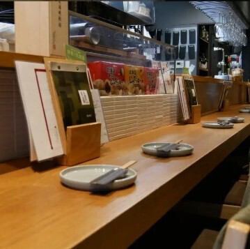 木の質感が心地よいカウンターに並ぶ器と箸。目の前で料理が仕上がるライブ感と、店主との距離の近さが、まるで家に帰ってきたような安心感を与えてくれる。肩肘張らず、気軽に美味しさと会話を楽しめる、そんな温もりある空間です