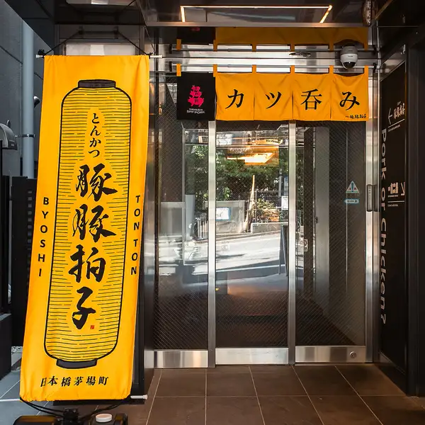 鮮やかなオレンジ色の旗が目印の入口。活気あふれる色彩が、店内の温かさと元気な雰囲気を予感させます。初めての方でも迷わずたどり着ける、親しみやすく明るい入り口です。ここから、とんかつとお酒の楽しい時間が始まります。