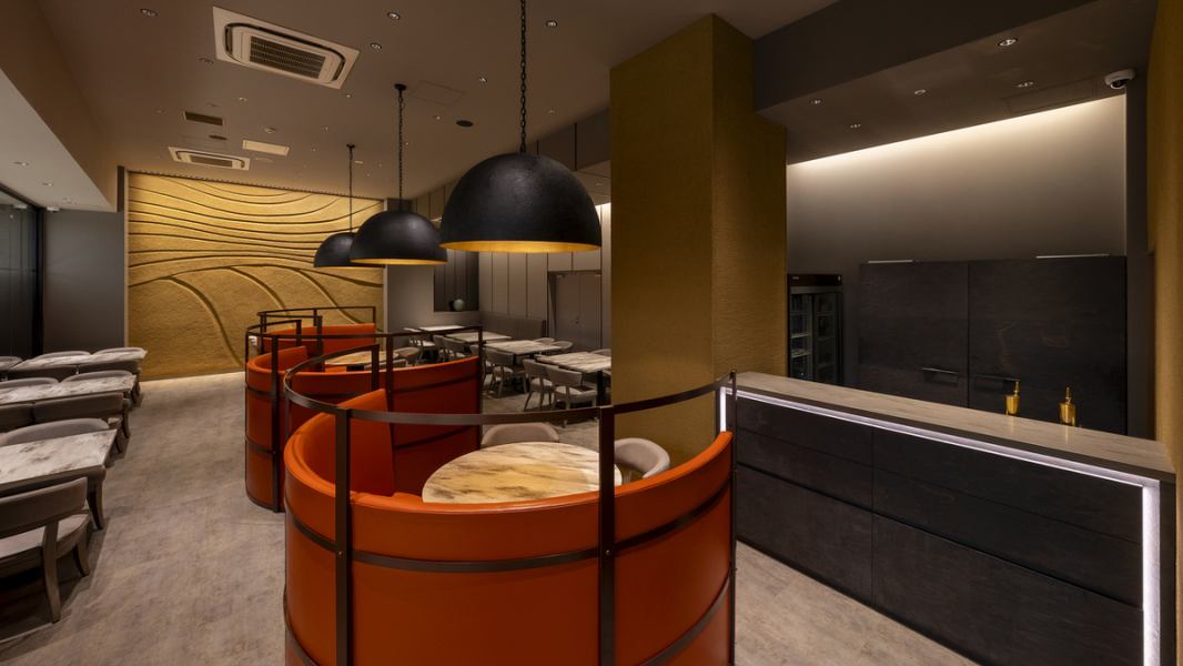 柔らかなオレンジ色のソファとシンプルなテーブルが配置され、リラックスできる雰囲気を醸し出しています。落ち着いたトーンの照明が、温かみを添え、食事や会話を楽しむのに最適な環境を提供。壁面のテクスチャーやディテールが、スタイリッシュな印象を与え、訪れる人々を魅了します。