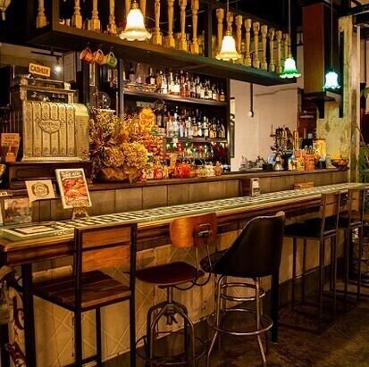 【カウンター席：6席】色とりどりのボトルが並ぶ本格的なバーカウンターは、まるで海外のダイナーのような雰囲気。バーテンダーとの会話を楽しみながら、自家製ジンジャーエールを使ったハイボールやモヒートなど、種類豊富なドリンクを味わえます。お一人様は勿論、デートやサク飲みにもぴったりな居心地の良い特等席です