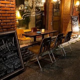 【テラス席】お店の顔である赤いレンガ壁を背景に、気軽に過ごせるテラス席をご用意しています。朝の光や夜のムードある照明の下で、開放的なひとときをお楽しみいただけます。モーニングやランチのカフェ利用はもちろん、ディナータイムには通りを眺めながら食事やカクテルを楽しむ、リラックスした時間をお過ごしください