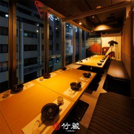 ［16～20名様：半個室］大きな窓から望む夜の街並みに、粋な和の意匠が彩りを添える団体様向けの半個室。木目のテーブルと落ち着いた照明が、静けさと温もりを感じさせ、ご宴席にふさわしい空間です。