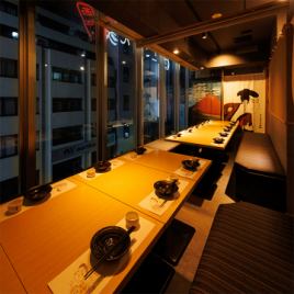 ［16～20名様：半個室］大きな窓から望む夜の街並みに、粋な和の意匠が彩りを添える団体様向けの半個室。木目のテーブルと落ち着いた照明が、静けさと温もりを感じさせ、ご宴席にふさわしい空間です。