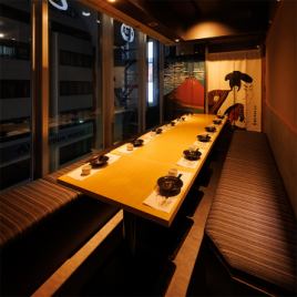 ［10～12名様：半個室］木の温もり感じる長テーブルと端正に設えた座席が、落着きを湛える和の半個室。窓から望む外の景色と、奥に掲げられた趣きある絵柄の仕切りが、非日常の情緒を添える特別席です。