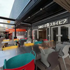 A rooftop beer garden bathed in midday sun.The open space created by the wide-open retractable tent is adorned with colorful lanterns, chairs, and neon decorations, creating a summery atmosphere.This is an extraordinary space where you can relax and enjoy daytime drinks or a BBQ while taking in the atmosphere of Umeda.
