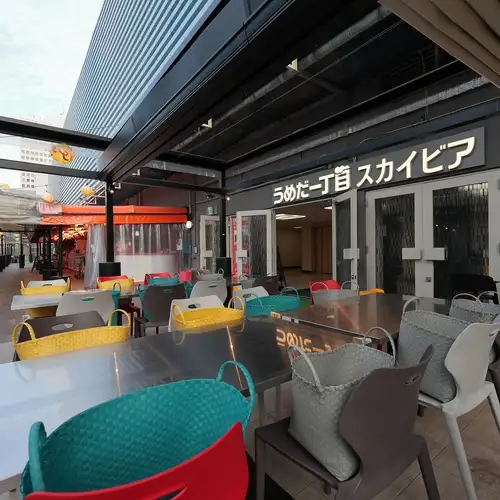 A rooftop beer garden bathed in midday sun.The open space created by the wide-open retractable tent is adorned with colorful lanterns, chairs, and neon decorations, creating a summery atmosphere.This is an extraordinary space where you can relax and enjoy daytime drinks or a BBQ while taking in the atmosphere of Umeda.